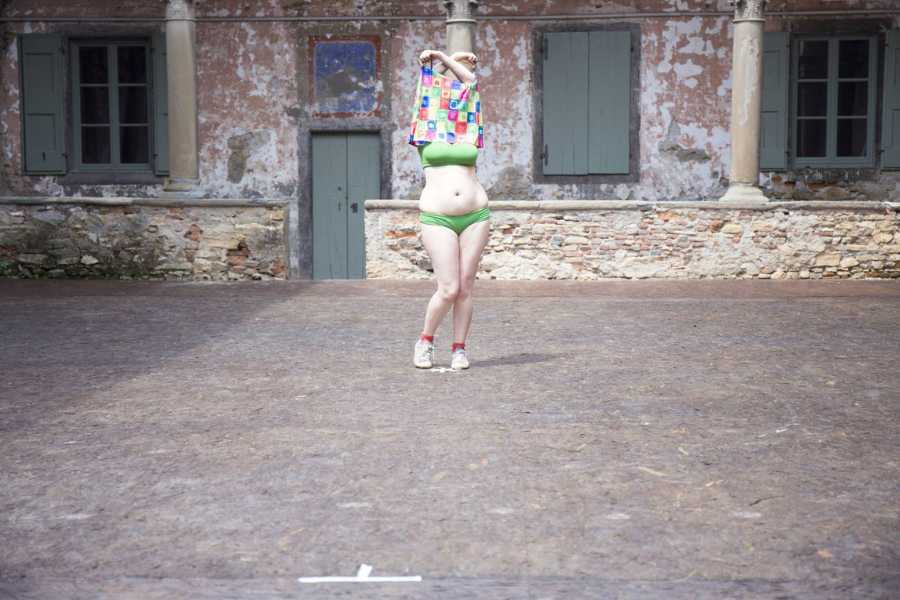 Woman stands in a courtyard lifting her brightly coloured dress over her head, revealing a green bikini