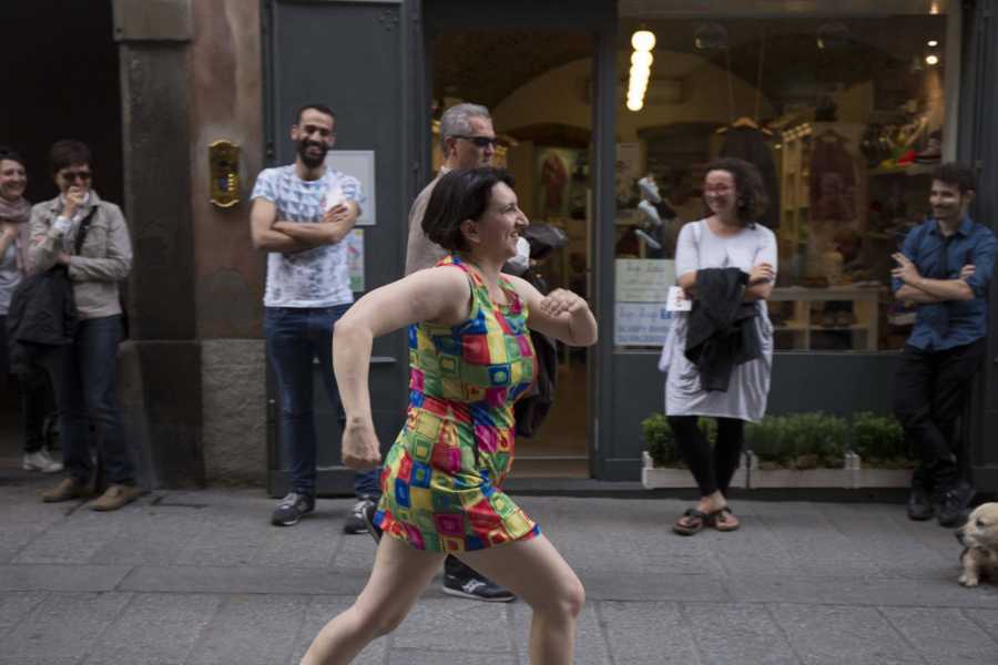 woman in a brightly coloured short dress runs down a street in front of storefronts as passersby look on