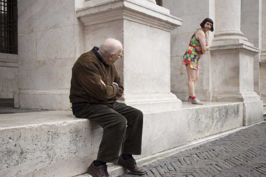 Woman in a brightly coloured short dress stands between outdoor pillars and peers at an elderly man seated on the steps