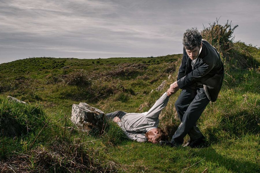 Outside in a field, one person drags another lifeless figure on the ground