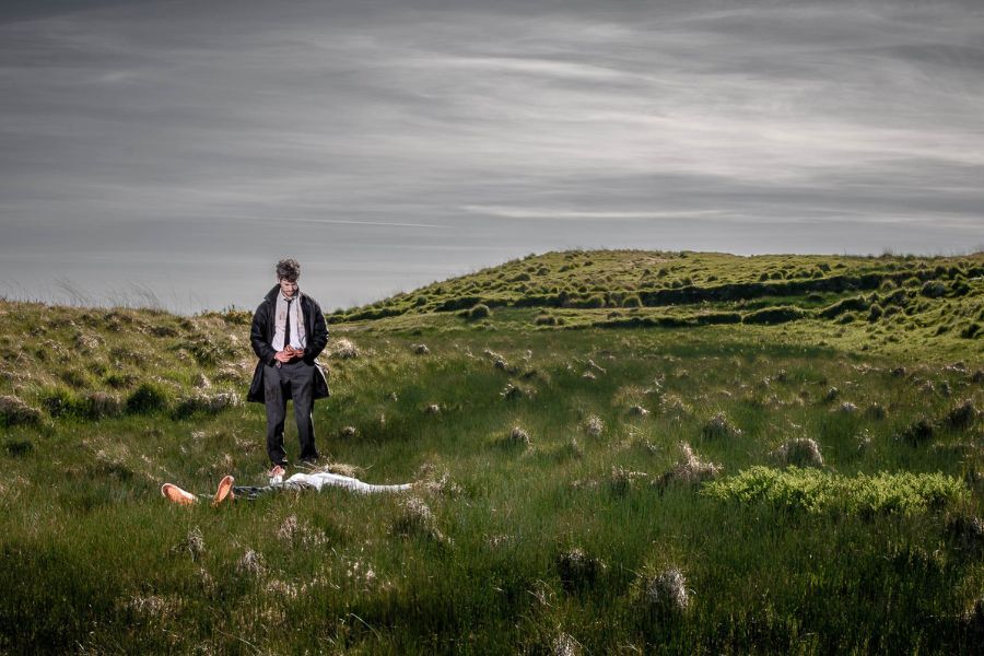 A figure lies largely obscured in a wide open field while another stands over him