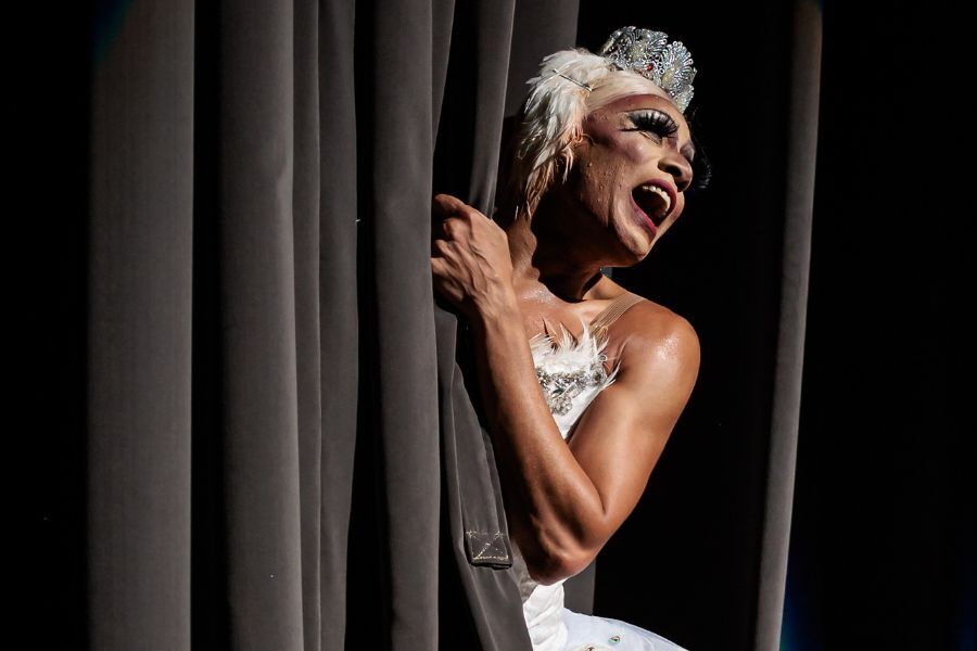 A male ballerina peeps out for the stage curtains and laughs with a melodramatic flair