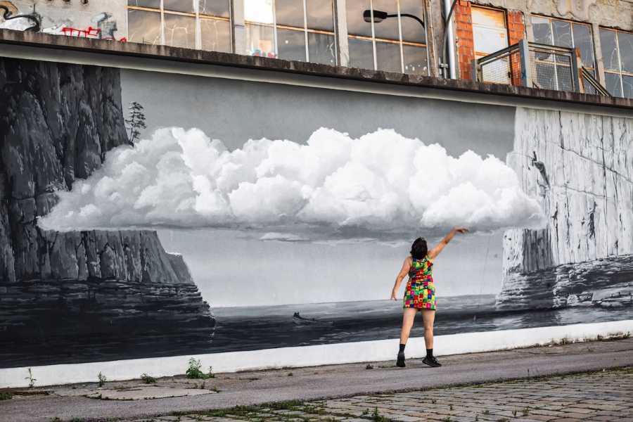 woman in a bright short dress stands in front of a graffiti wall lifting her hand as if to touch the painted cloud