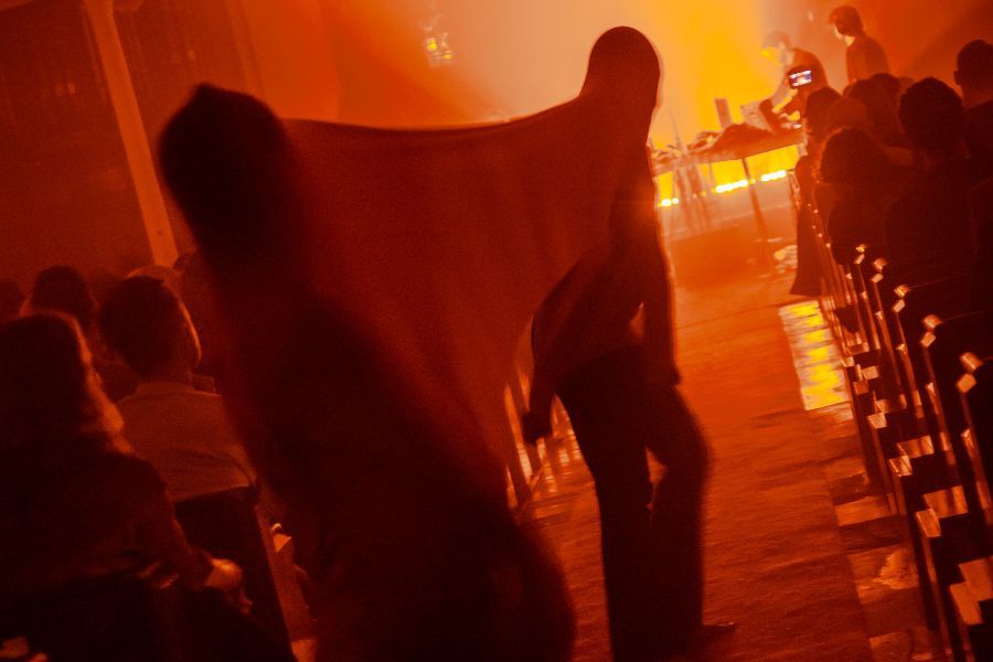 A hazy scene in orange lighting. Two figures with fabric stretch over their heads are silhouetted and dance down the aisle as people are seated in the benches on either side.