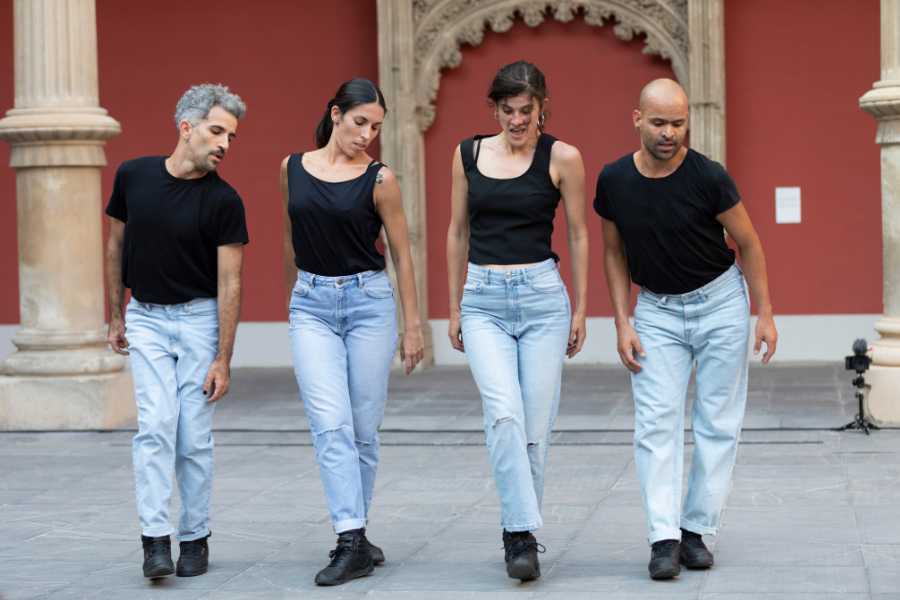 Four people in jeans and black tops perform in unison outdoors in front of a red coloured building