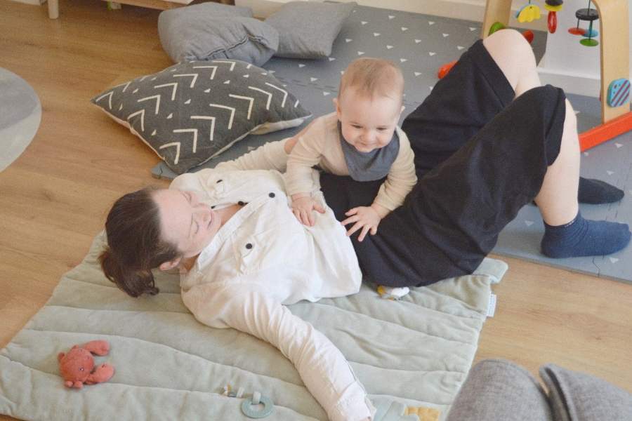 A woman lies on her back on the floor while her baby crawls on her tummy