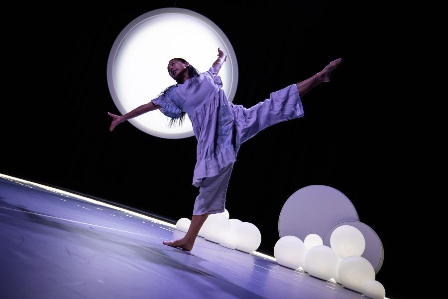 A dancer leans back on one leg with the other high in the air and arms outstretched. A full moon glows in the background.