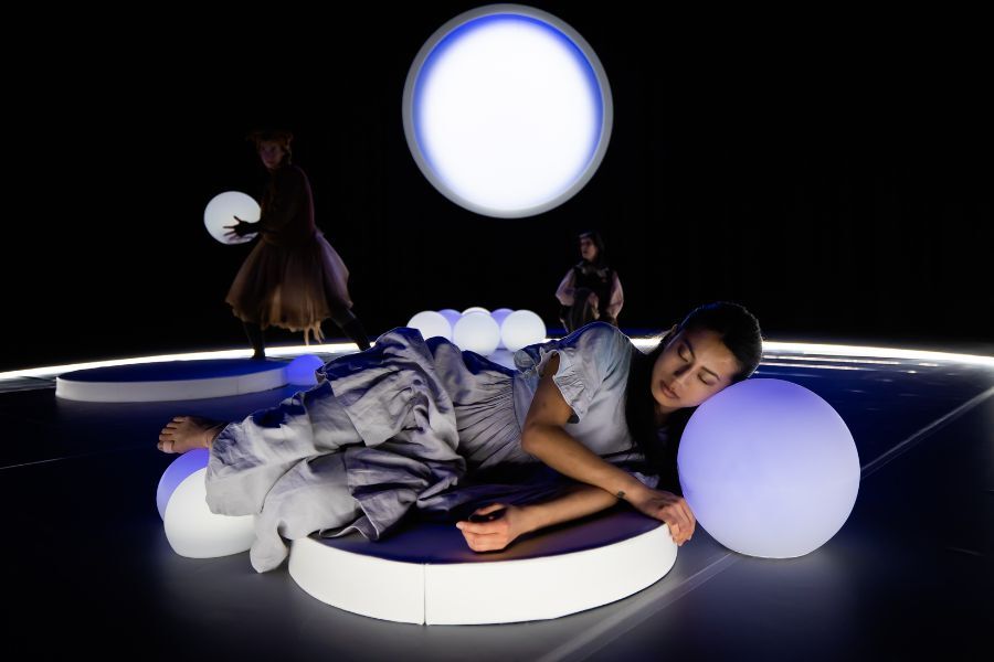 On stage, a performers dressed in white lies with her head on a glowing globe with another shining in the background
