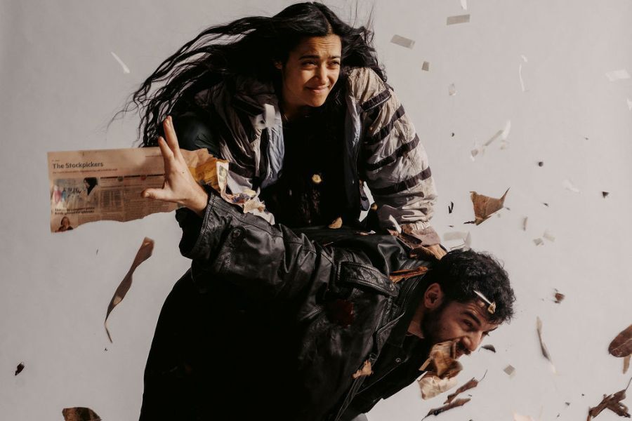 One figure holds another on his back while they face strong wind and debris blows all around them