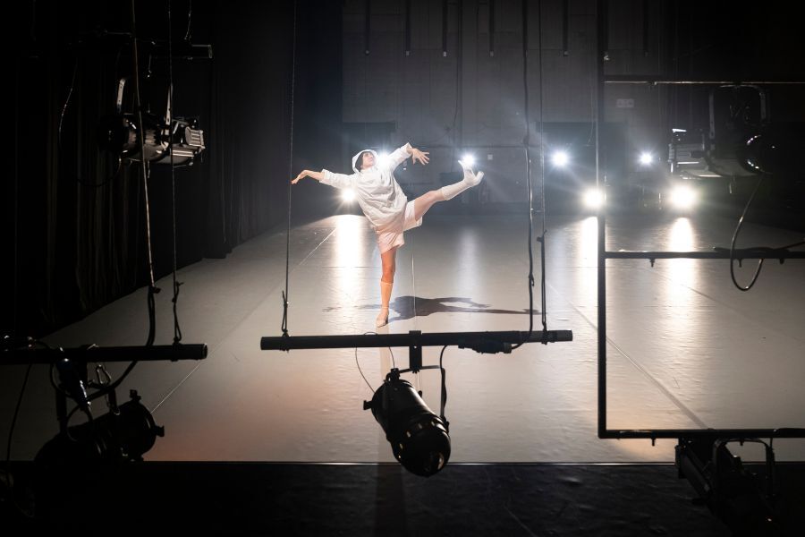 One dancer on a dark stage illuminated by spotlights and wearing white shorts and a hoodie leans to the side and elevates her leg high