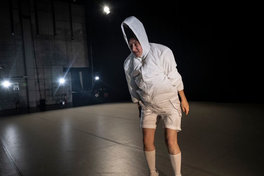 One dancer on a dark stage wearing white shorts and a hoodie leans over and looks at the ground. Her hood is on her head and stretch up as if something is on her head underneath the hood.