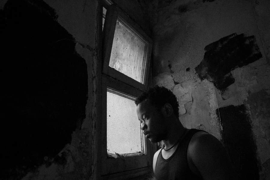 A man sits in a start room at the window, his face gently illuminated by the outdoor light.