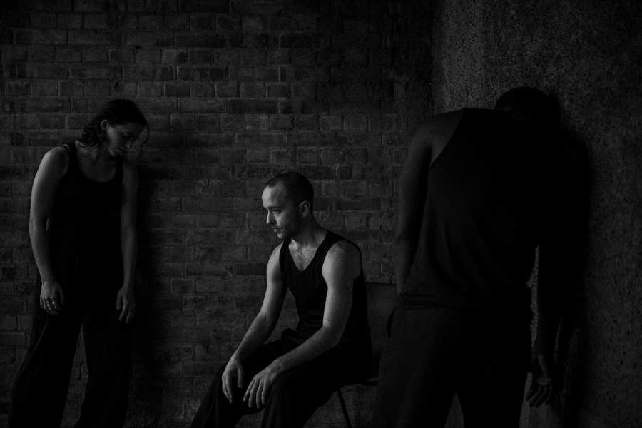 Two figures in a dimly lit bare room, the man sits on a chair and the woman stands nearby looking downwards.