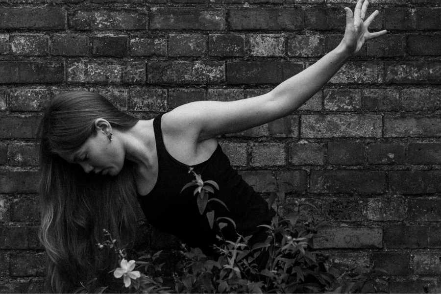 Black and white image of a woman in front of a brick wall looking down to the side and lifting her one arm up behind her.