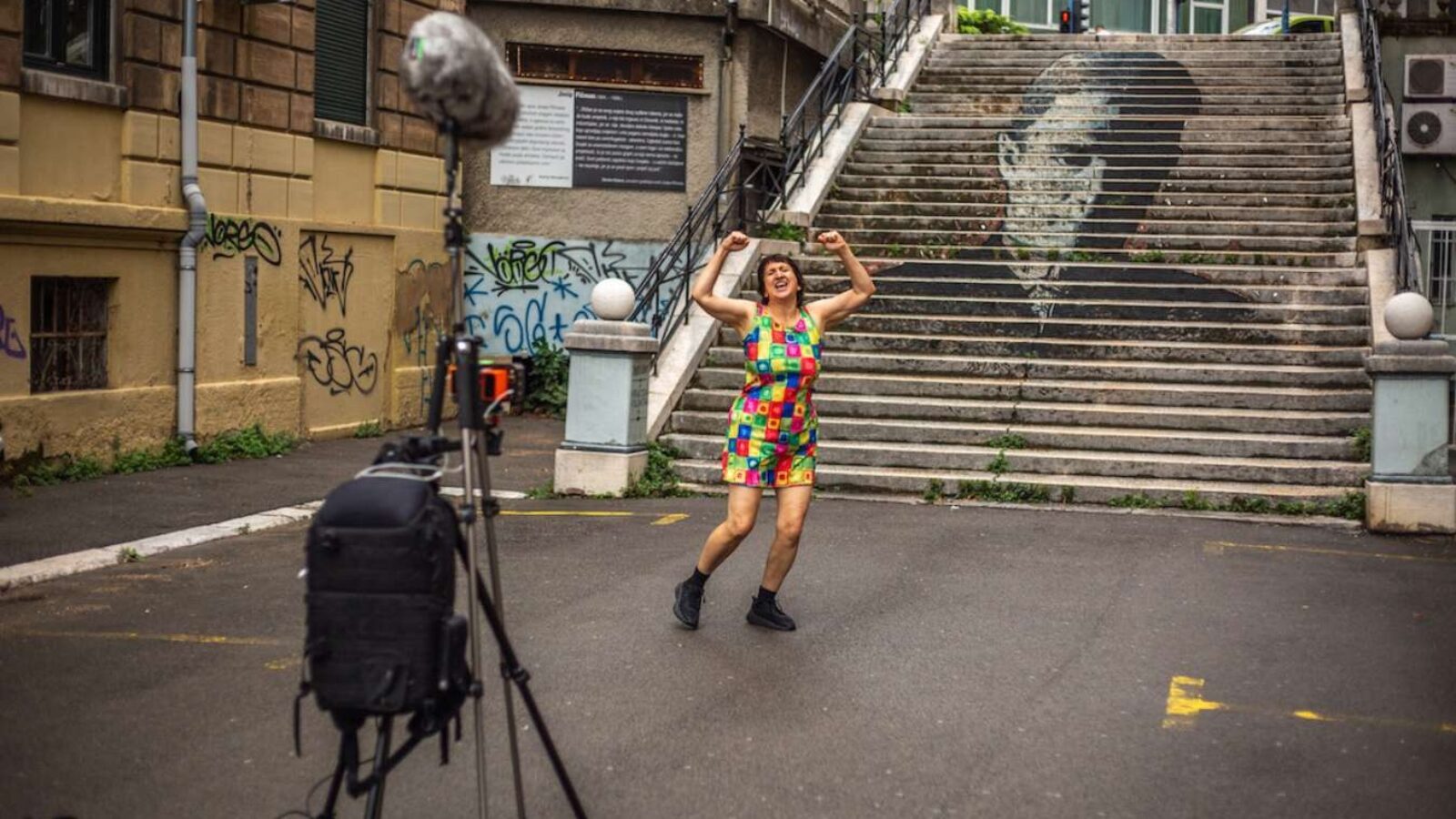 A woman in a brightly coloured jumpsuit stands at the base of outdoor steps with a filming setup in front