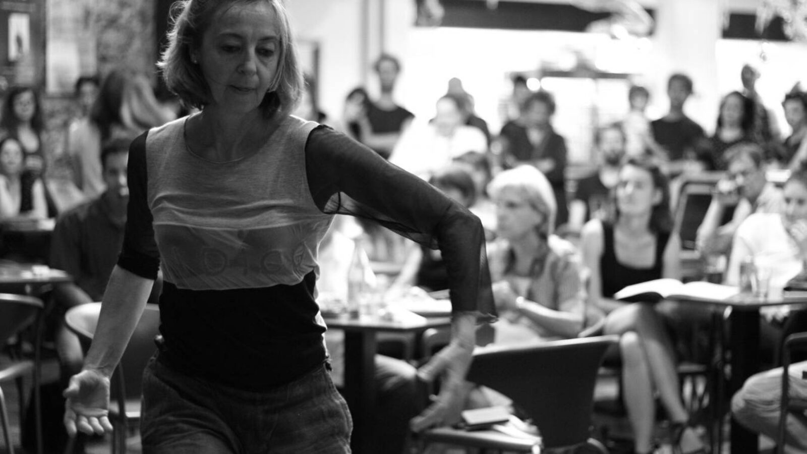 A black and while photo of a woman dancing in the foreground with people seated at small round tables in the background.