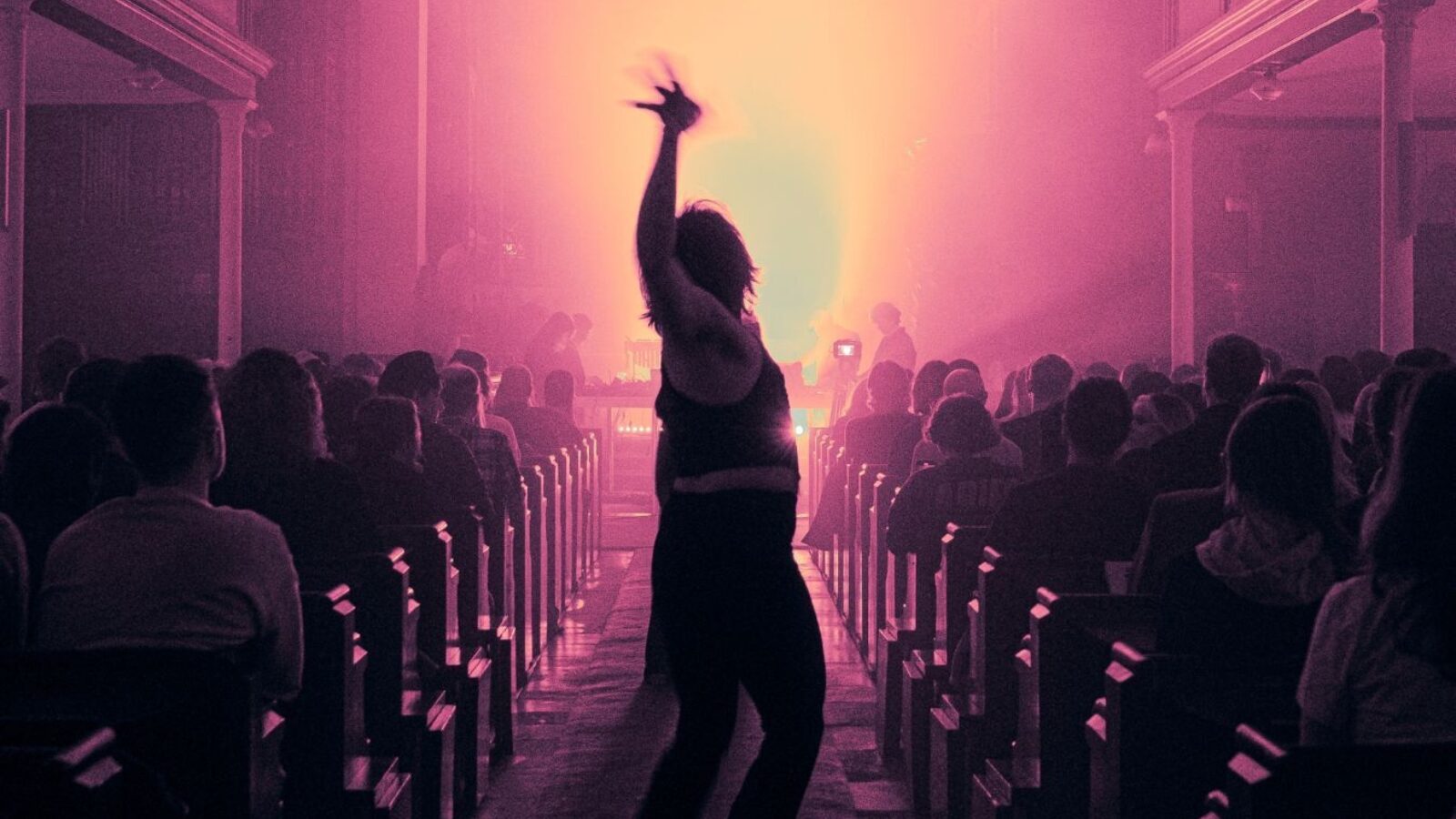 A hazy scene in pink lighting. A single figure silhouetted dances down the aisle as people are seated in the benches on either side.