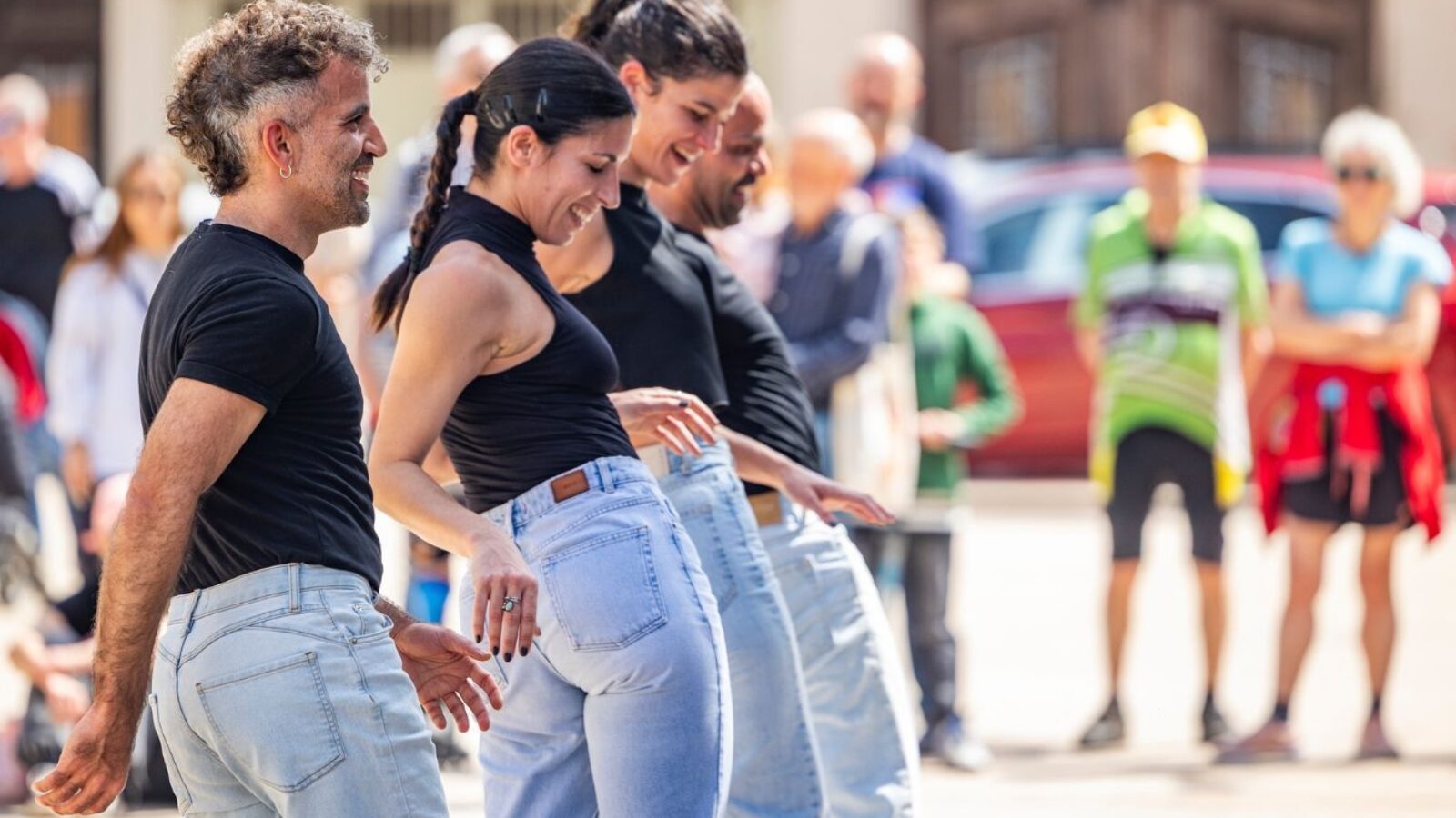 Four people in jeans and black tops perform in unison outdoors
