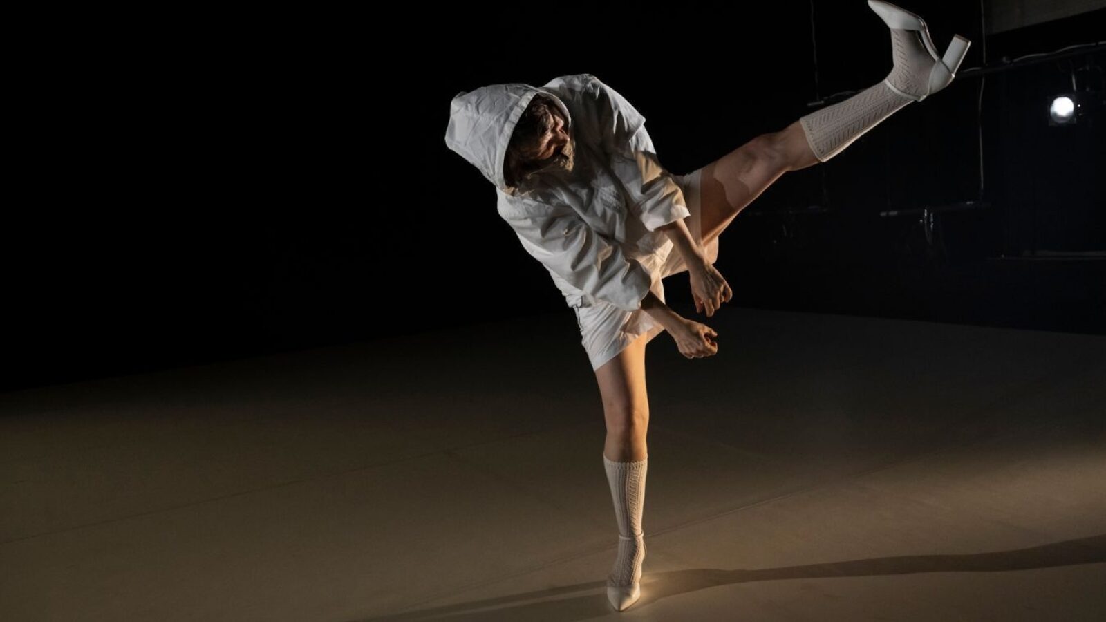 One dancer on a dark stage wearing white shorts and a hoodie leans to the side and elevates her leg high