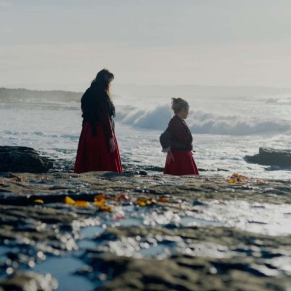 Two women wade in the shallows of the sea with the sun glinting off the rocks in the foreground
