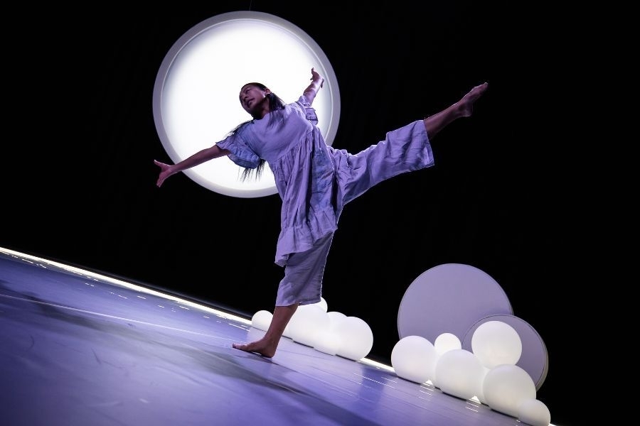 A dancer leans back on one leg with the other high in the air and arms outstretched. A full moon glows in the background.