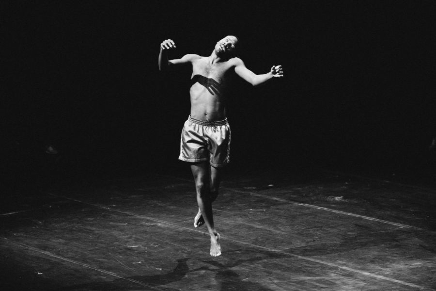 A man dancing on a plain stage, wearing only green coloured shorts.