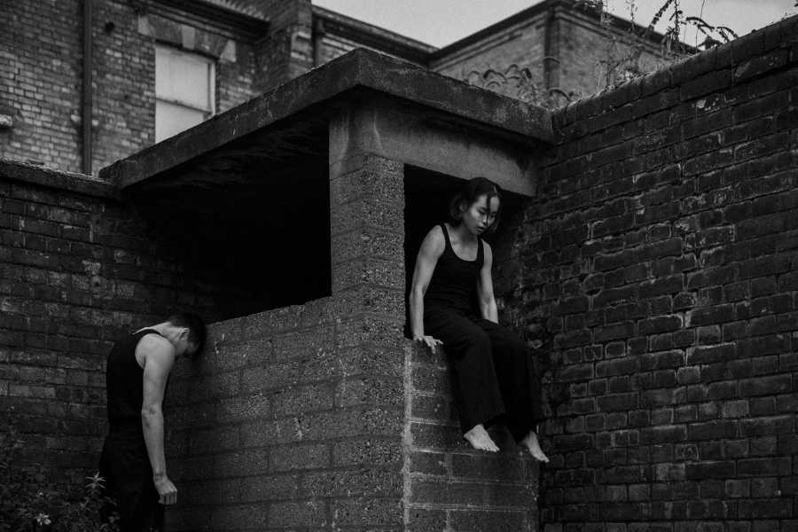 Two figures in a bricked courtyard. The man stands and leans his head against the wall, the woman sits in a window space, dangling her legs and looking down.