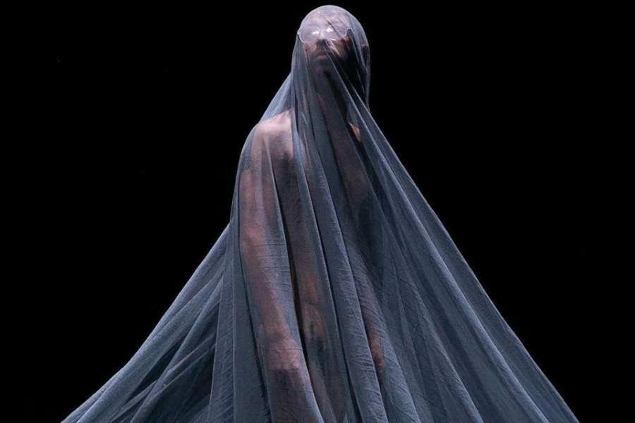 A single standing figure draped in sheer fabric against a black background