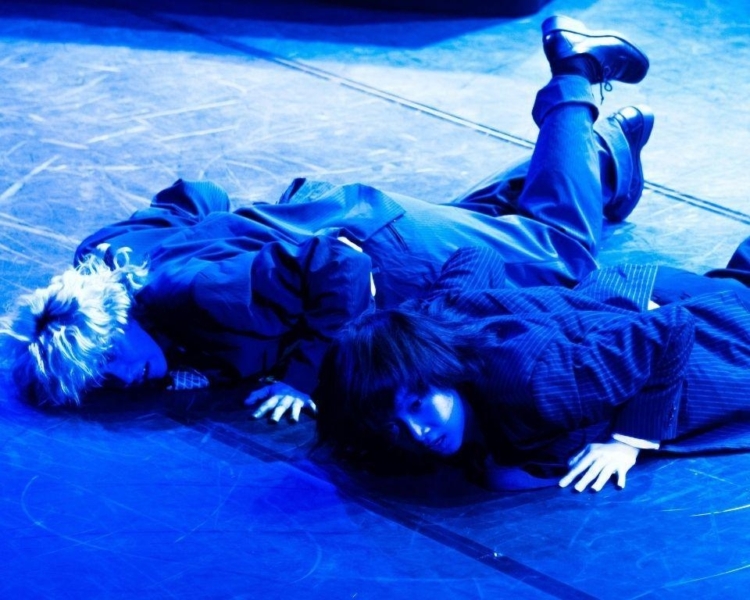 Two performer lie on their stomachs on the floor, bathed in blue light