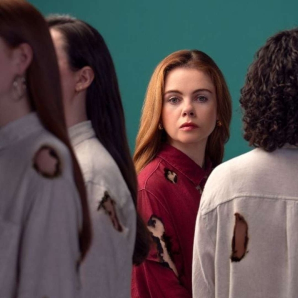 Woman wearing a red shirt burnt by fire looks at us past figures in similar burnt clothes in the foreground