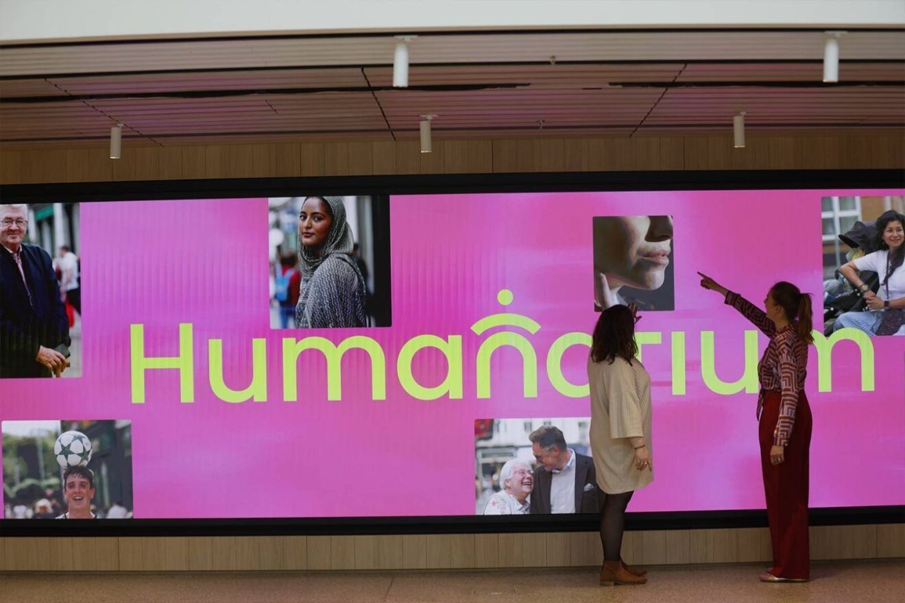 Two people looking at a large format digital screen displaying a bright pink background with portrait photos of various people and the word 'Humanarium'
