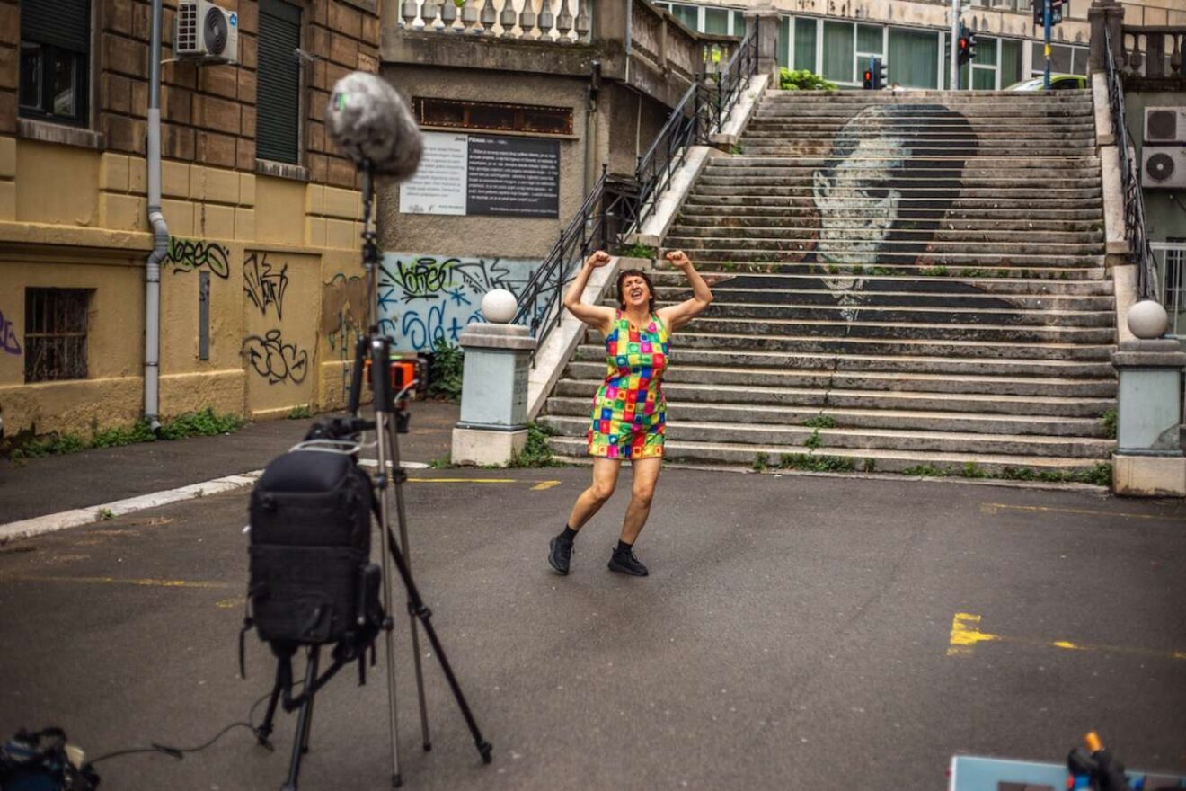 A woman in a brightly coloured jumpsuit stands at the base of outdoor steps with a filming setup in front