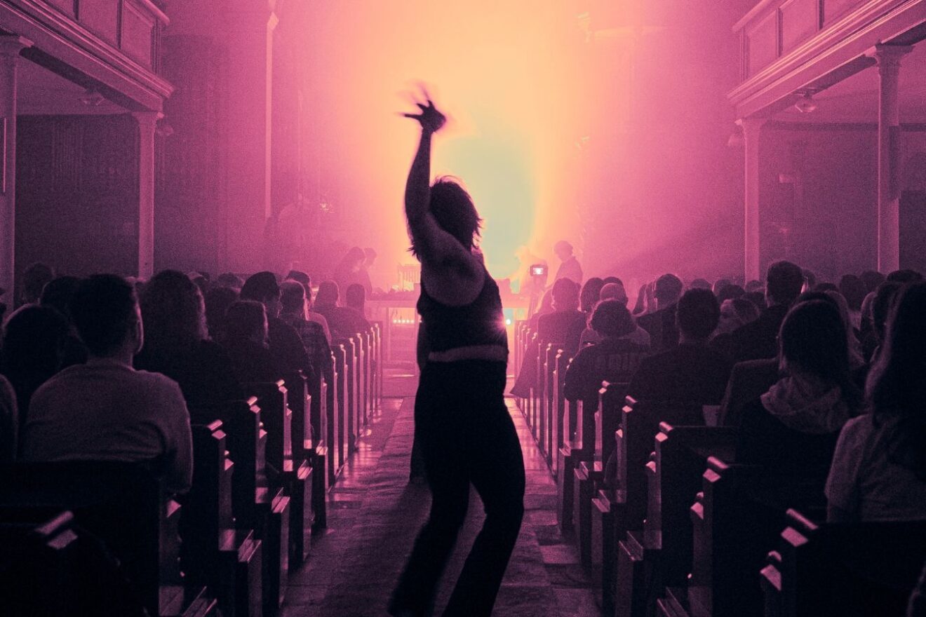 A hazy scene in pink lighting. A single figure silhouetted dances down the aisle as people are seated in the benches on either side.