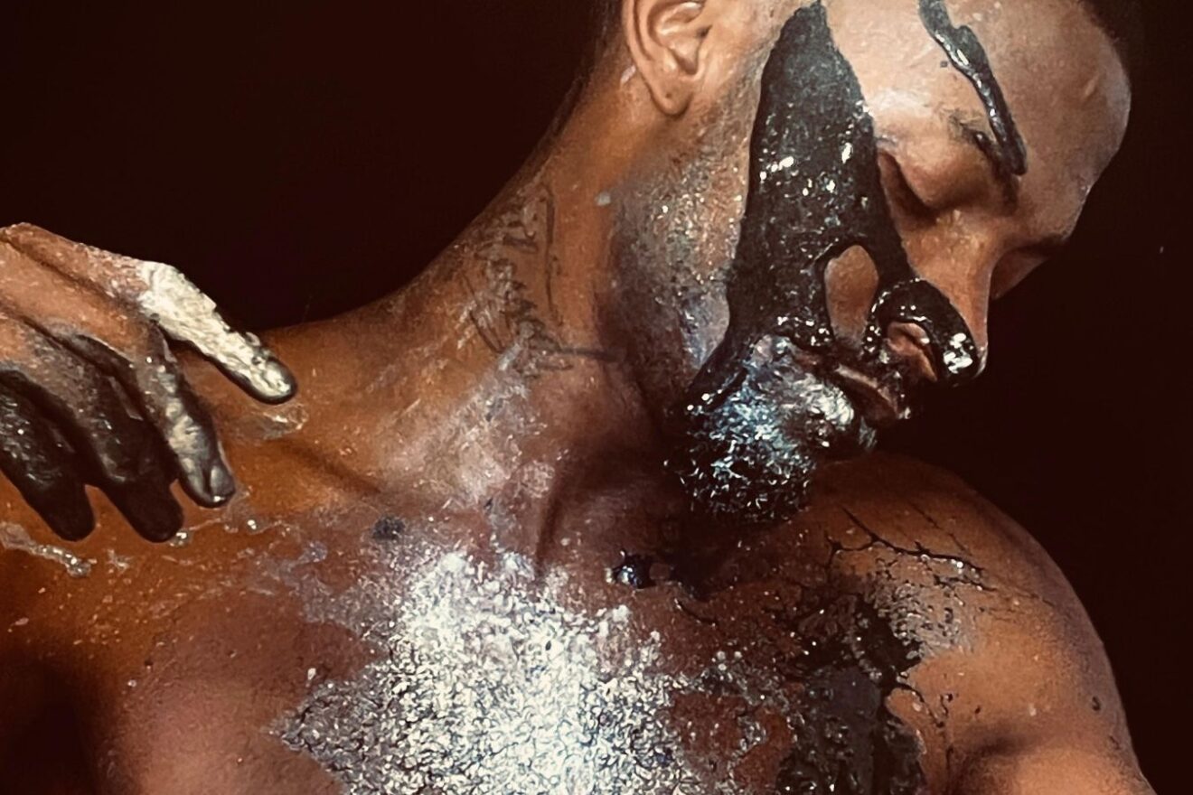 A man with dark skin streaked with shiny silver paint, shown from the showers up.