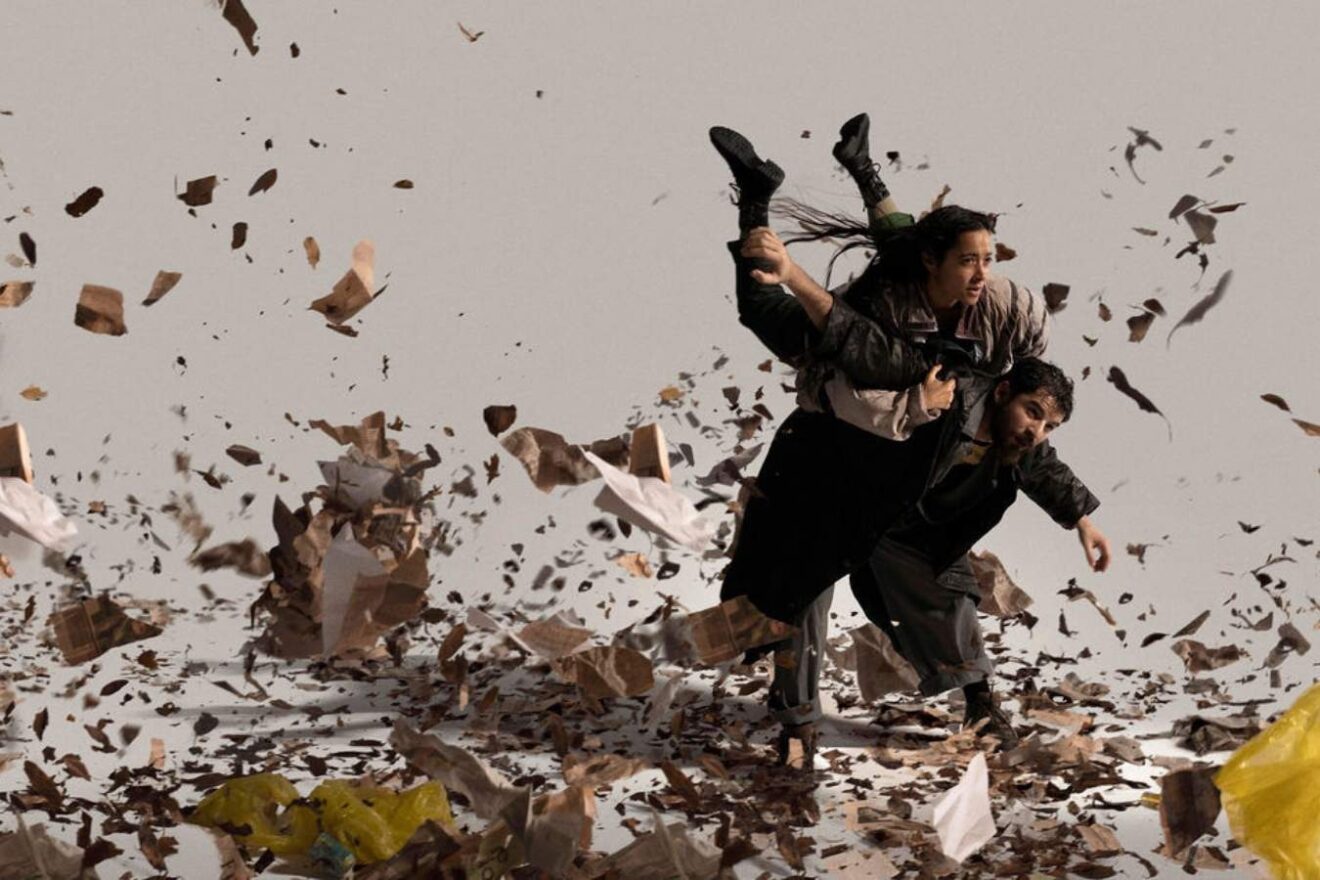 One figure holds another on his back while they face strong wind and debris blows all around them