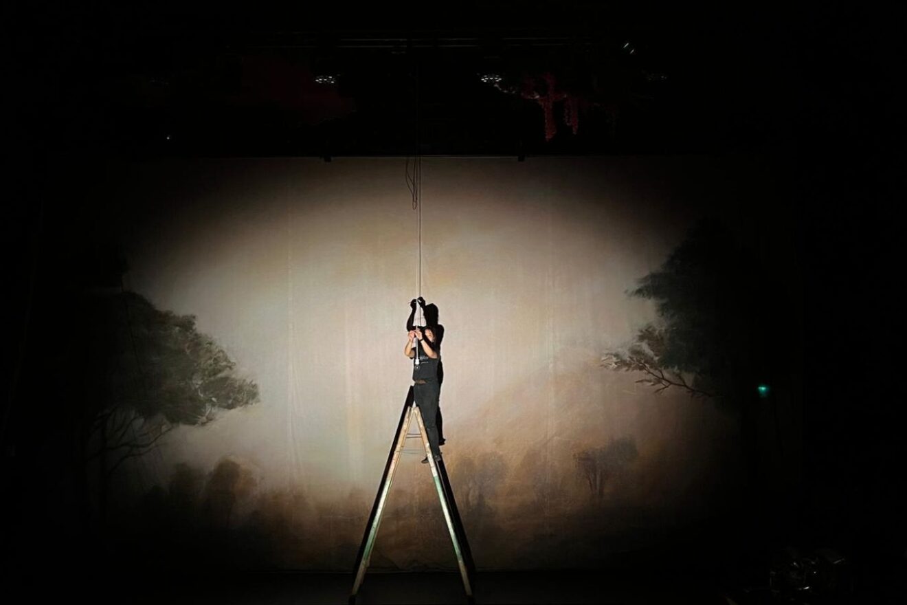 A dark stage with a spotlight on a person standing on a ladder.