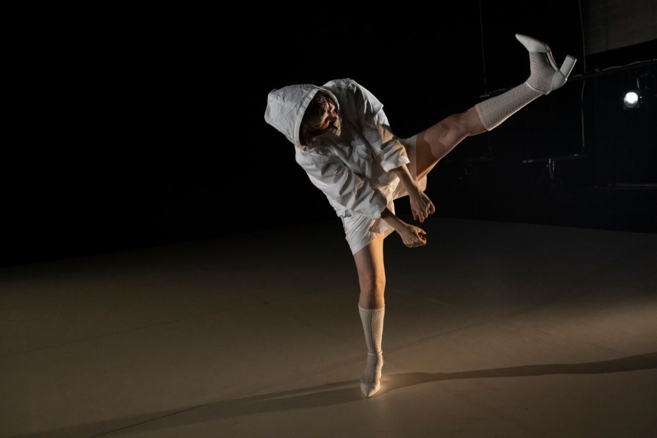 One dancer on a dark stage wearing white shorts and a hoodie leans to the side and elevates her leg high