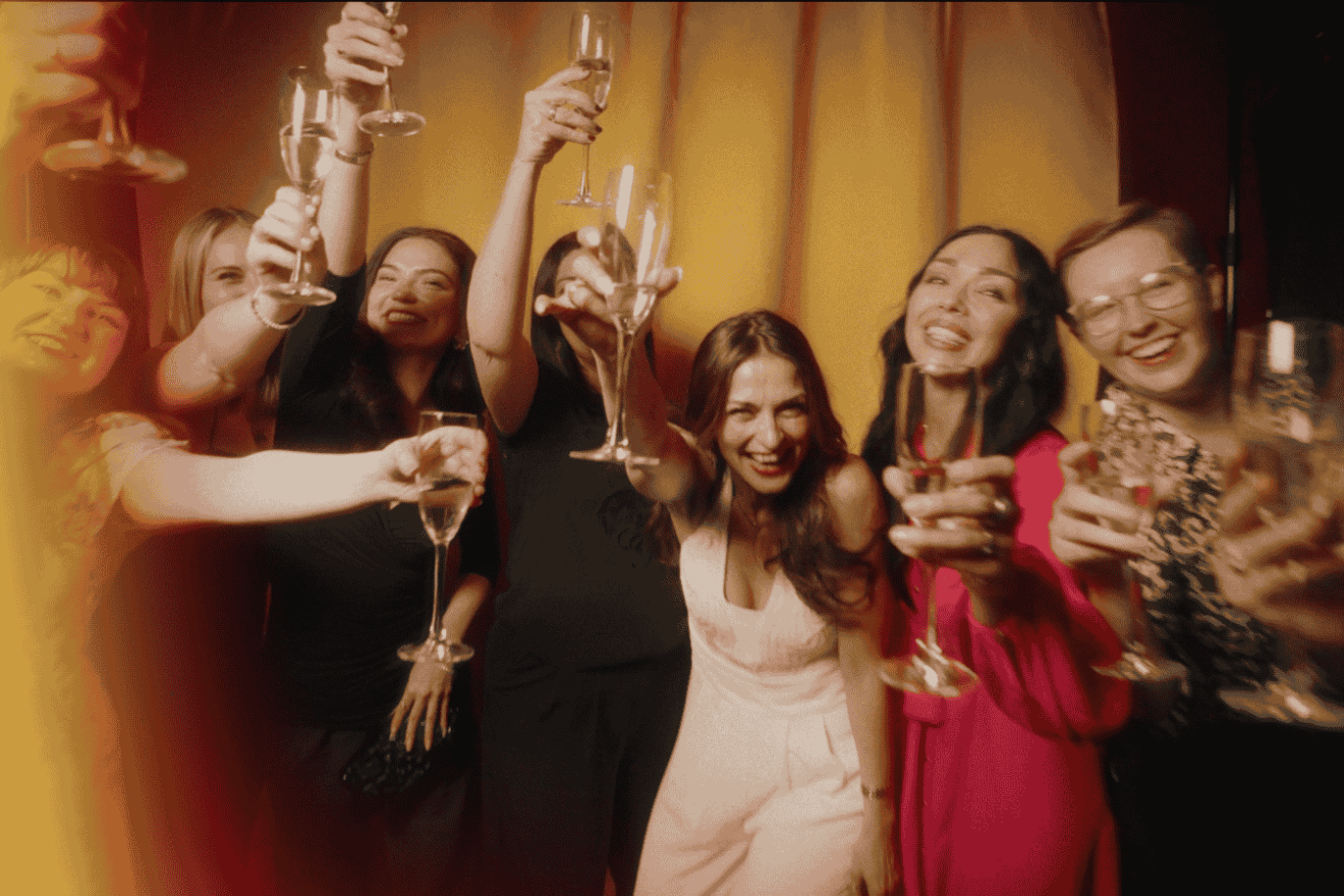 A joyful group raising their champagne glasses in a celebratory toast, smiling and leaning toward the camera against a warm, golden background.