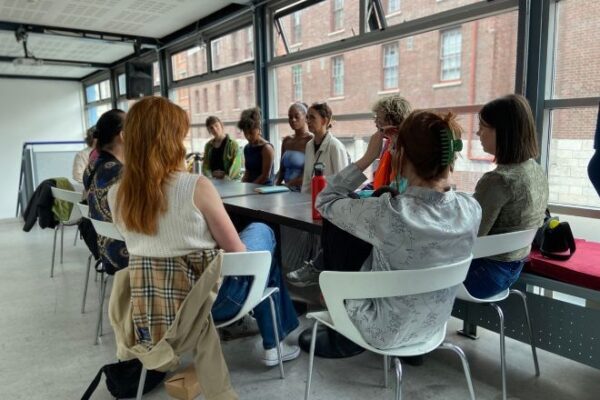 Group of dance artists sit around a table in discussion in a bright room with windows that run along the length of the room.