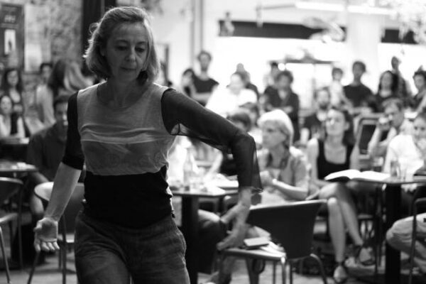 A black and while photo of a woman dancing in the foreground with people seated at small round tables in the background.