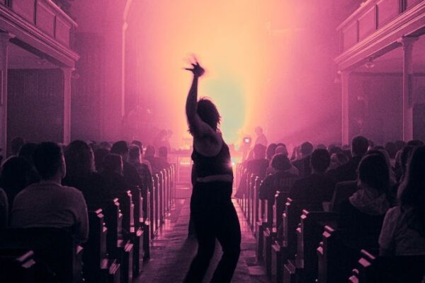 A hazy scene in pink lighting. A single figure silhouetted dances down the aisle as people are seated in the benches on either side.