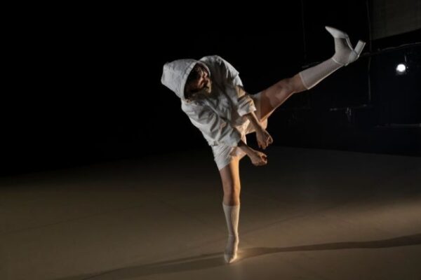 One dancer on a dark stage wearing white shorts and a hoodie leans to the side and elevates her leg high