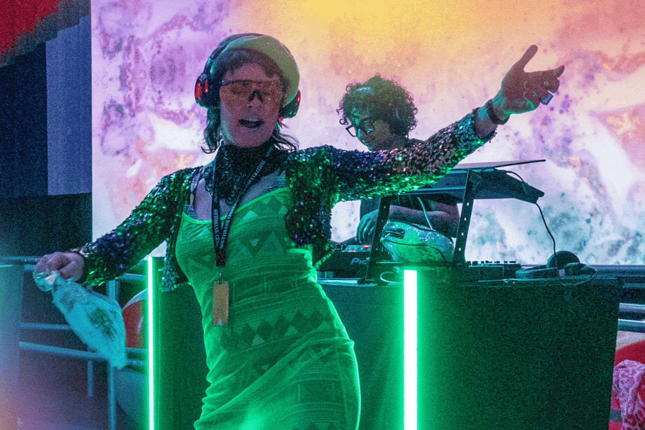 A person in a bright green geometric dress and sequin jacket dancing enthusiastically at a silent disco event, wearing headphones and tinted glasses, with a DJ performing in the background under colourful, psychedelic lighting.